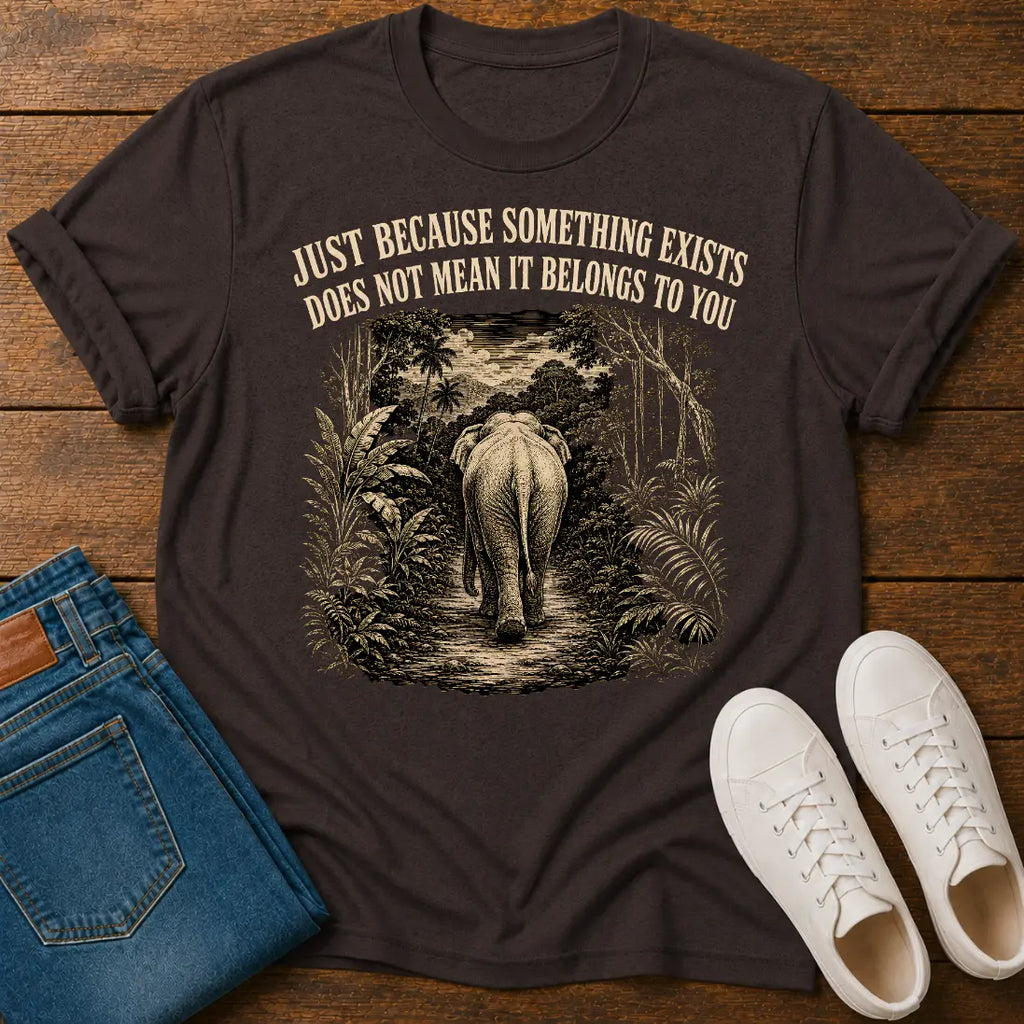 A dark brown graphic t-shirt laid flat on a wooden surface, styled with blue jeans and white sneakers. The t-shirt features a graphic of an elephant walking through a jungle path with the text 'JUST BECAUSE SOMETHING EXISTS DOES NOT MEAN IT BELONGS TO YOU'.