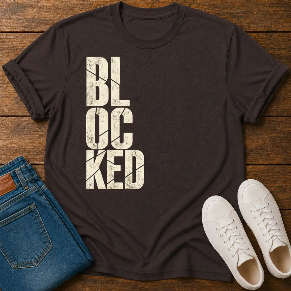 A dark brown t-shirt with a distressed cream-colored 'BLOCKED' graphic print, styled in a flat lay on a wooden background alongside blue jeans and white sneakers.