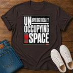 A dark brown graphic t-shirt laid flat on a wooden surface, styled with folded blue jeans and a pair of white sneakers. The t-shirt features a bold graphic print that reads 'UNAPOLOGETICALLY OCCUPING SPACE' in white and red text inside a black square.