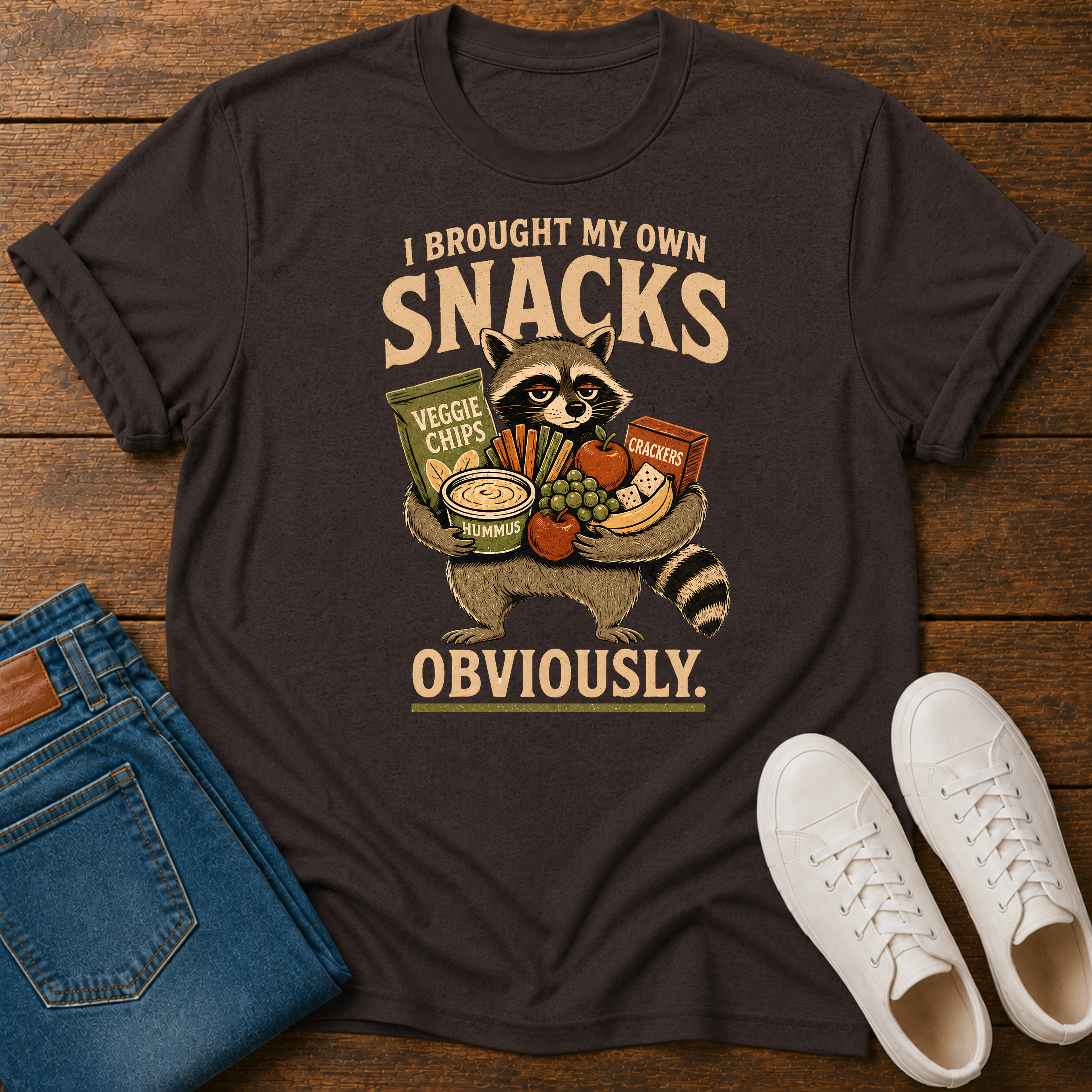A dark brown graphic t-shirt laid flat on a wooden surface, styled with blue jeans and white sneakers. The t-shirt features a cartoon raccoon holding various snacks with the text 'I BROUGHT MY OWN SNACKS OBVIOUSLY.