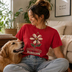 A woman sitting on a beige couch in a cozy living room, wearing a red graphic t-shirt with a flower design and the text 'COMPASSION REBELLION'. A golden retriever dog is sitting next to her.