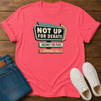 A coral pink graphic t-shirt laid flat on a dark wood background, styled with folded blue jeans and white sneakers. The t-shirt features a retro marquee-style graphic that reads 'NOT UP FOR DEBATE' with 'VACANCY FOR PEACE' and 'OPINIONS FULL' underneath.