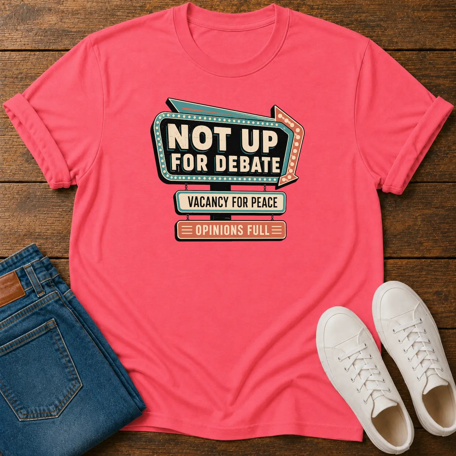 A coral pink graphic t-shirt laid flat on a dark wood background, styled with folded blue jeans and white sneakers. The t-shirt features a retro marquee-style graphic that reads 'NOT UP FOR DEBATE' with 'VACANCY FOR PEACE' and 'OPINIONS FULL' underneath.