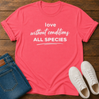 A coral pink short-sleeve t-shirt with white text reading 'love without conditions ALL SPECIES' laid flat on a dark wood background, styled with folded blue jeans and white sneakers.
