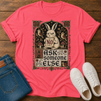 A coral pink graphic t-shirt laid flat on a rustic wooden background, styled with folded blue jeans and white sneakers. The t-shirt features a vintage-style illustration of a rabbit holding a sign that says 'NO' surrounded by birds and mushrooms, with the text 'ASK someone ELSE' below.