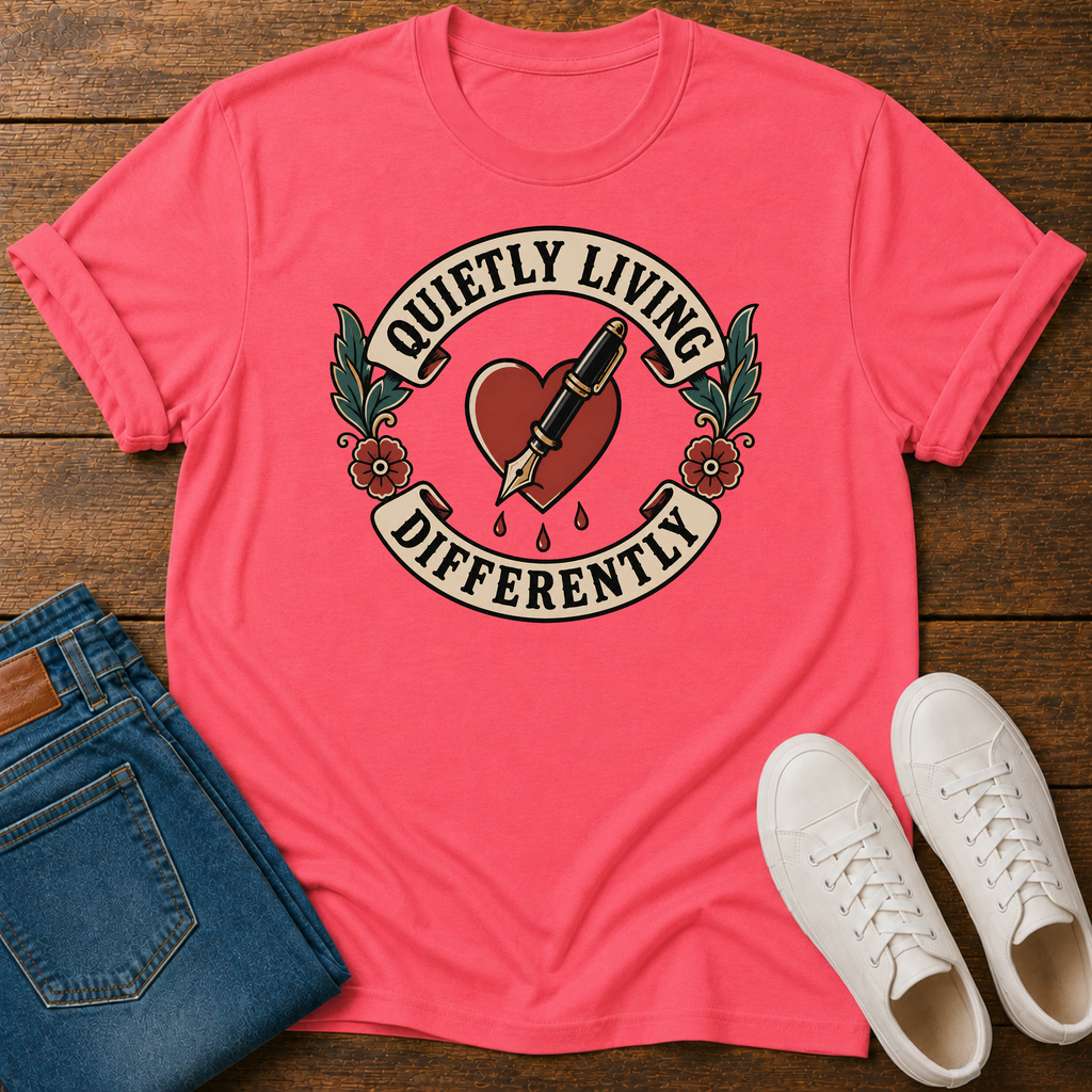 A coral pink graphic t-shirt laid flat on a dark wood background, styled with folded blue jeans and white sneakers. The t-shirt features a tattoo-style graphic of a red heart with a fountain pen nib, green leaves, and a cream banner reading 'QUIETLY LIVING DIFFERENTLY'.