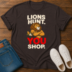 A dark brown graphic t-shirt laid flat on a wooden surface, styled with folded blue jeans and white sneakers. The t-shirt features a graphic of a lion with crossed arms holding a shopping cart, with the text 'LIONS HUNT.YOU SHOP'