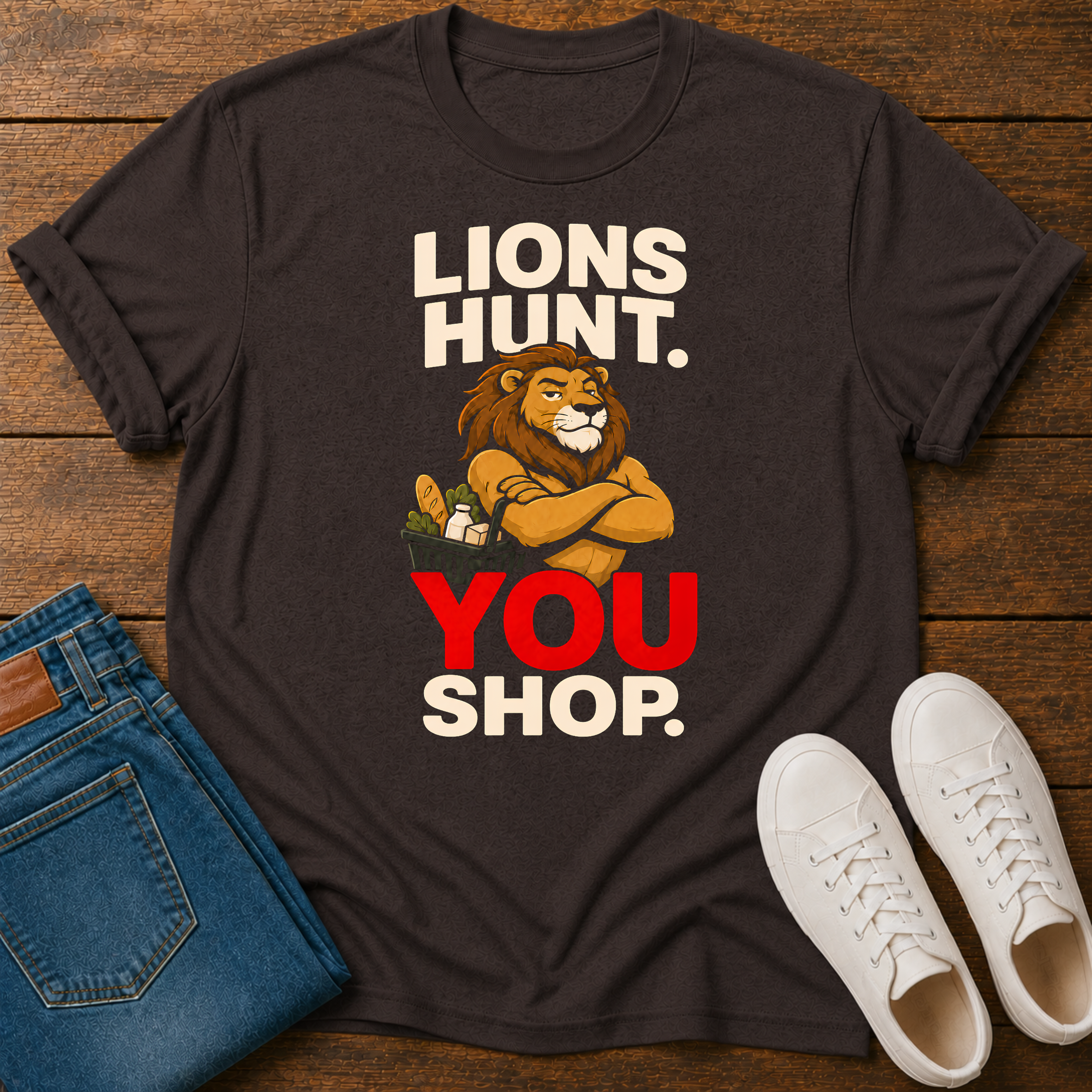 A dark brown graphic t-shirt laid flat on a wooden surface, styled with folded blue jeans and white sneakers. The t-shirt features a graphic of a lion with crossed arms holding a shopping cart, with the text 'LIONS HUNT.YOU SHOP'