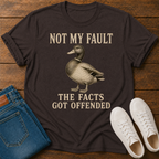 A dark brown graphic t-shirt laid flat on a wooden surface, styled with blue jeans and white sneakers. The t-shirt features a vintage-style illustration of a mallard duck and the text 'NOT MY FAULT THE FACTS GOT OFFENDED' in a beige serif font.