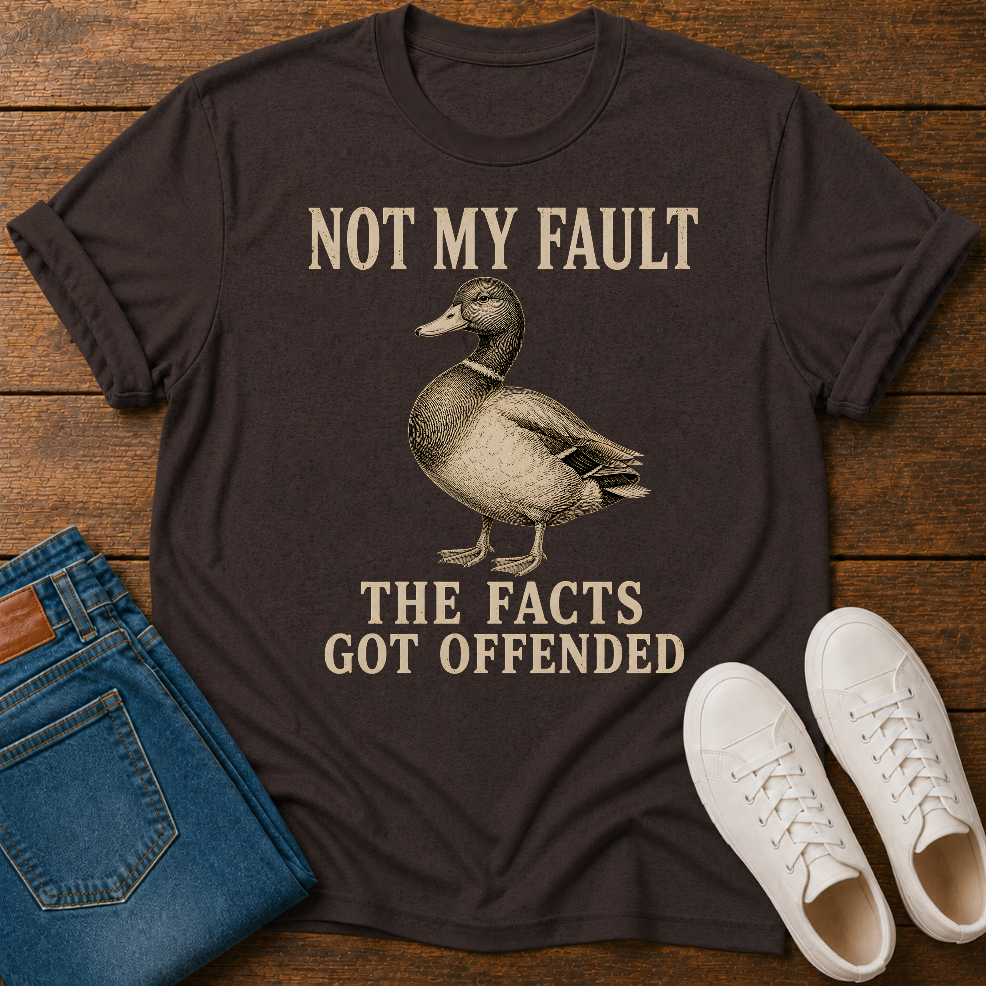 A dark brown graphic t-shirt laid flat on a wooden surface, styled with blue jeans and white sneakers. The t-shirt features a vintage-style illustration of a mallard duck and the text 'NOT MY FAULT THE FACTS GOT OFFENDED' in a beige serif font.