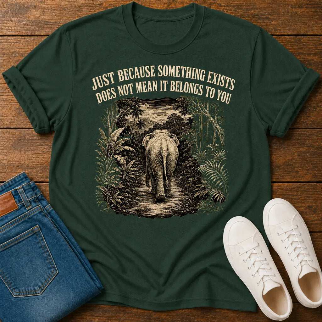 A dark green graphic t-shirt laid flat on a wooden surface, styled with folded blue jeans and white sneakers. The t-shirt features a vintage-style illustration of an elephant walking through a jungle, with the text 'JUST BECAUSE SOMETHING EXISTS DOES NOT MEAN IT BELONGS TO YOU' printed above the graphic.
