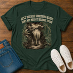 A dark green graphic t-shirt laid flat on a wooden surface, styled with folded blue jeans and white sneakers. The t-shirt features a vintage-style illustration of an elephant walking through a jungle, with the text 'JUST BECAUSE SOMETHING EXISTS DOES NOT MEAN IT BELONGS TO YOU' printed above the graphic.