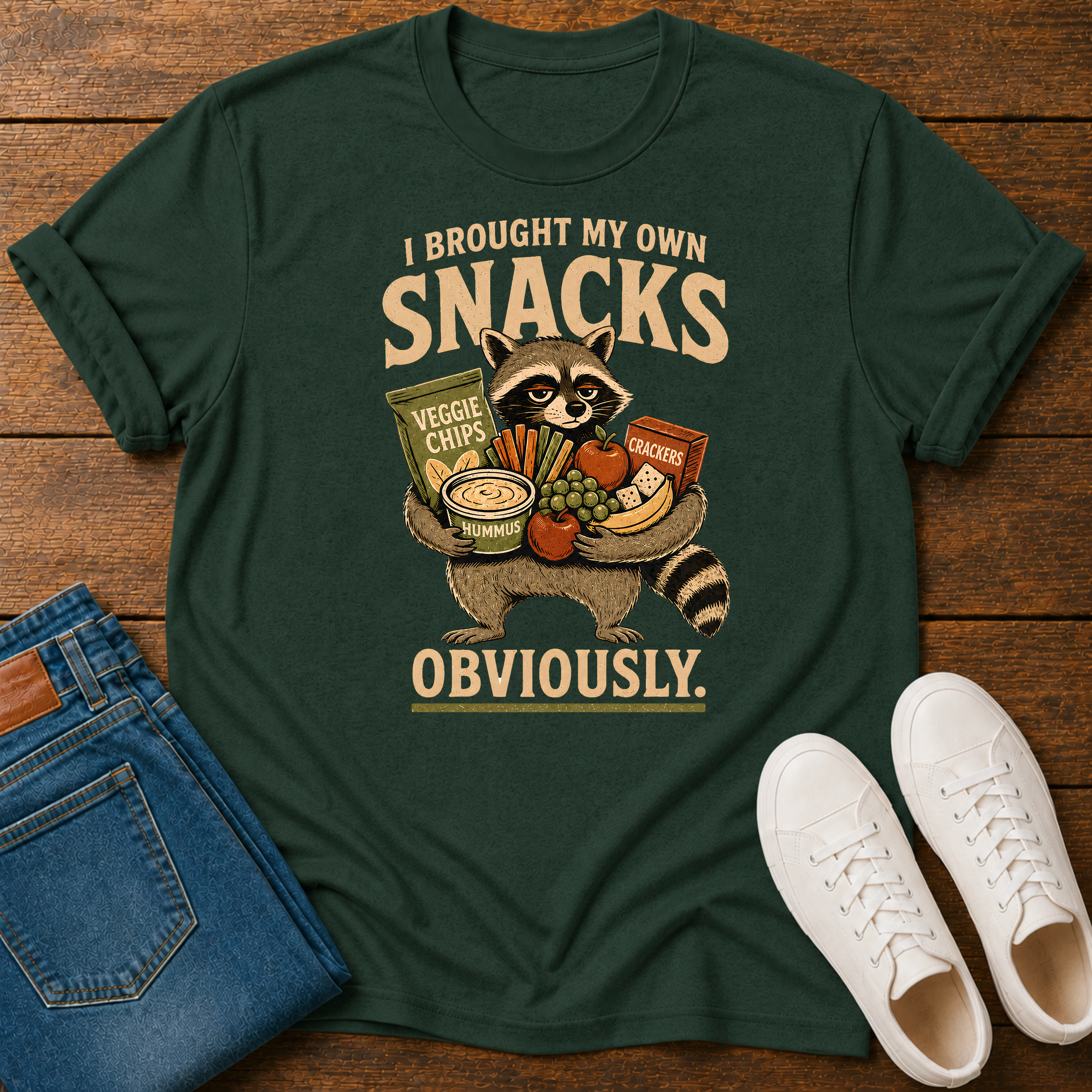A dark green graphic t-shirt laid flat on a wooden surface, styled with folded blue jeans and white sneakers. The t-shirt features a cartoon raccoon holding various snacks with the text 'I BROUGHT MY OWN SNACKS OBVIOUSLY.