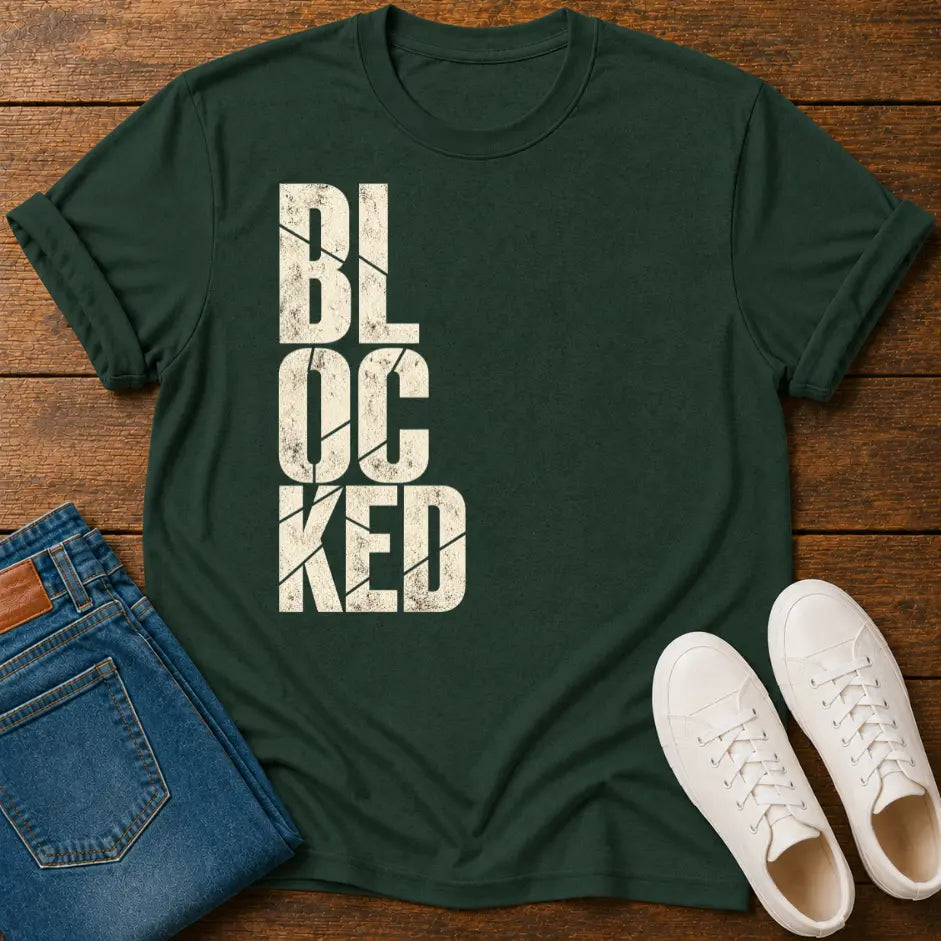 A dark green crewneck t-shirt with the word 'BLOCKED' printed vertically in distressed off-white block letters, styled in a flat lay on a wooden surface alongside folded blue jeans and a pair of white sneakers.