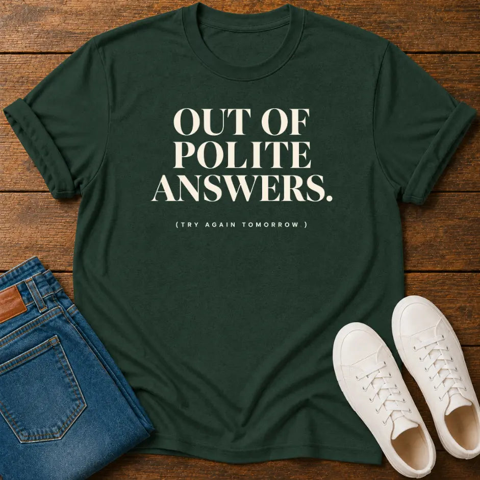 A dark green graphic t-shirt with the text 'OUT OF POLITE ANSWERS. (TRY AGAIN TOMORROW)' printed in white, styled in a flat-lay on a wooden background with blue jeans and white sneakers.