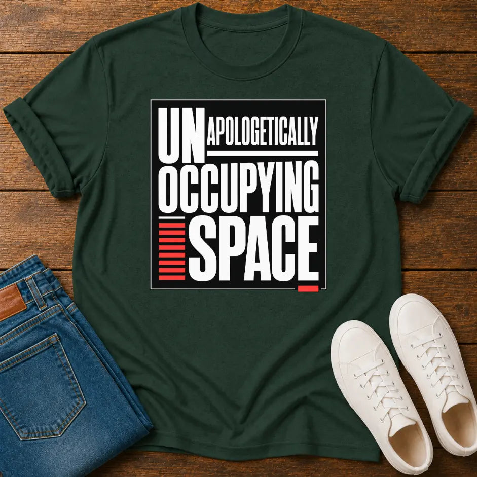 A dark green graphic t-shirt laid flat on a wooden surface, styled with folded blue jeans and white sneakers. The t-shirt features a bold white and red text graphic that reads 'UNAPOLOGETICALLY OCCUPYING SPACE'.