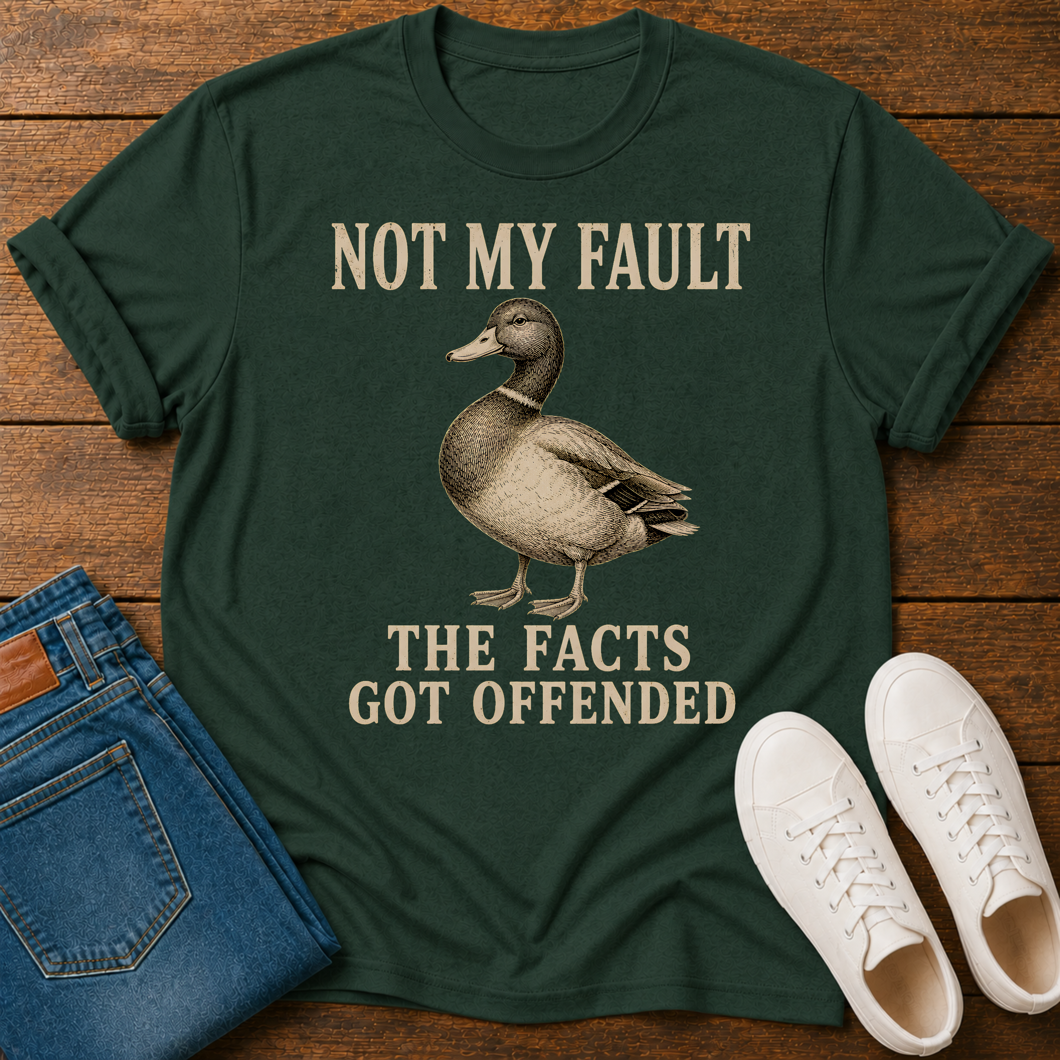 A dark green graphic t-shirt featuring a duck illustration and the text 'NOT MY FAULT THE FACTS GOT OFFENDED' in beige, styled in a flat-lay with blue jeans and white sneakers on a wooden background.