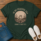 A dark green graphic t-shirt featuring a vintage-style frog illustration and the text 'IN MY PEACEFUL ERA (DON'T TEST IT)' in beige. The t-shirt is styled in a flat-lay arrangement on a wooden surface, accompanied by folded blue jeans and a pair of white sneakers.