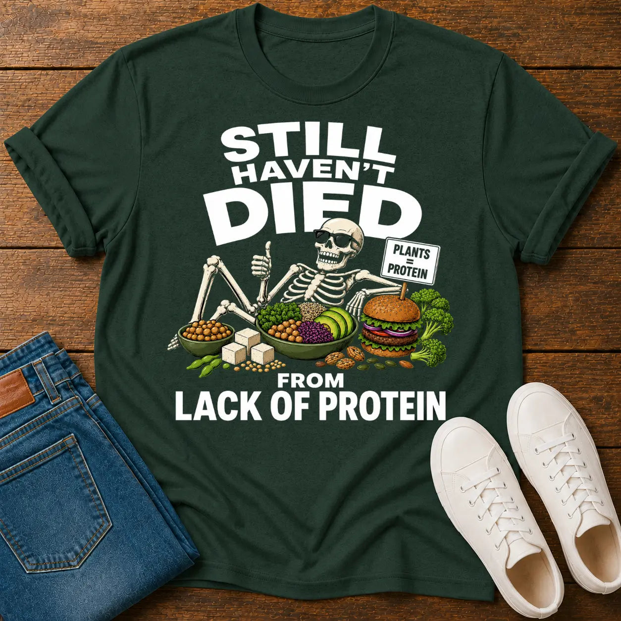 Green t-shirt with skeleton graphic and text on a wooden surface with jeans and shoes.