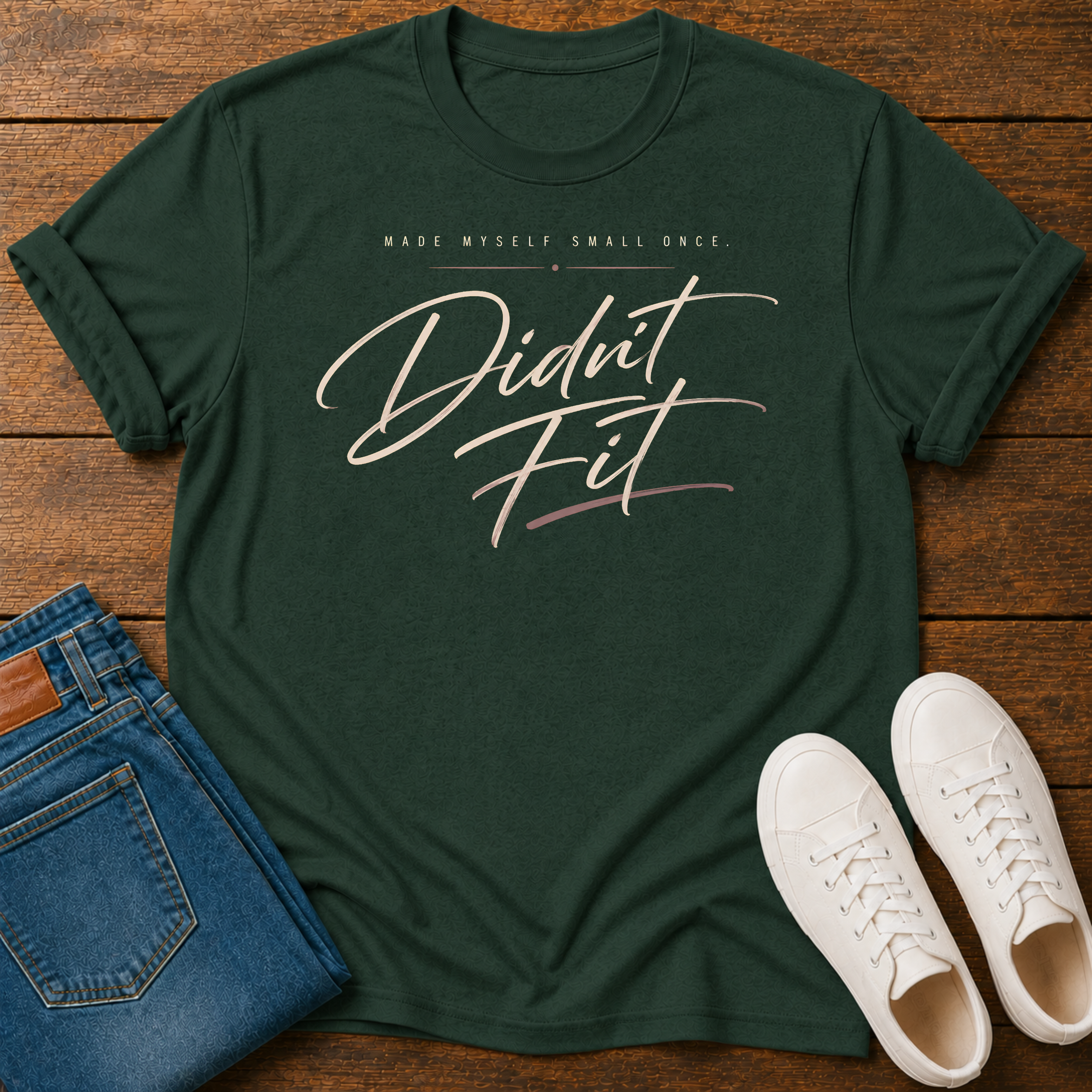 A dark green graphic t-shirt laid flat on a wooden surface, styled with folded blue jeans and white sneakers. The t-shirt features the text 'MADE MYSELF SMALL ONCE. DIDN'T FIT'