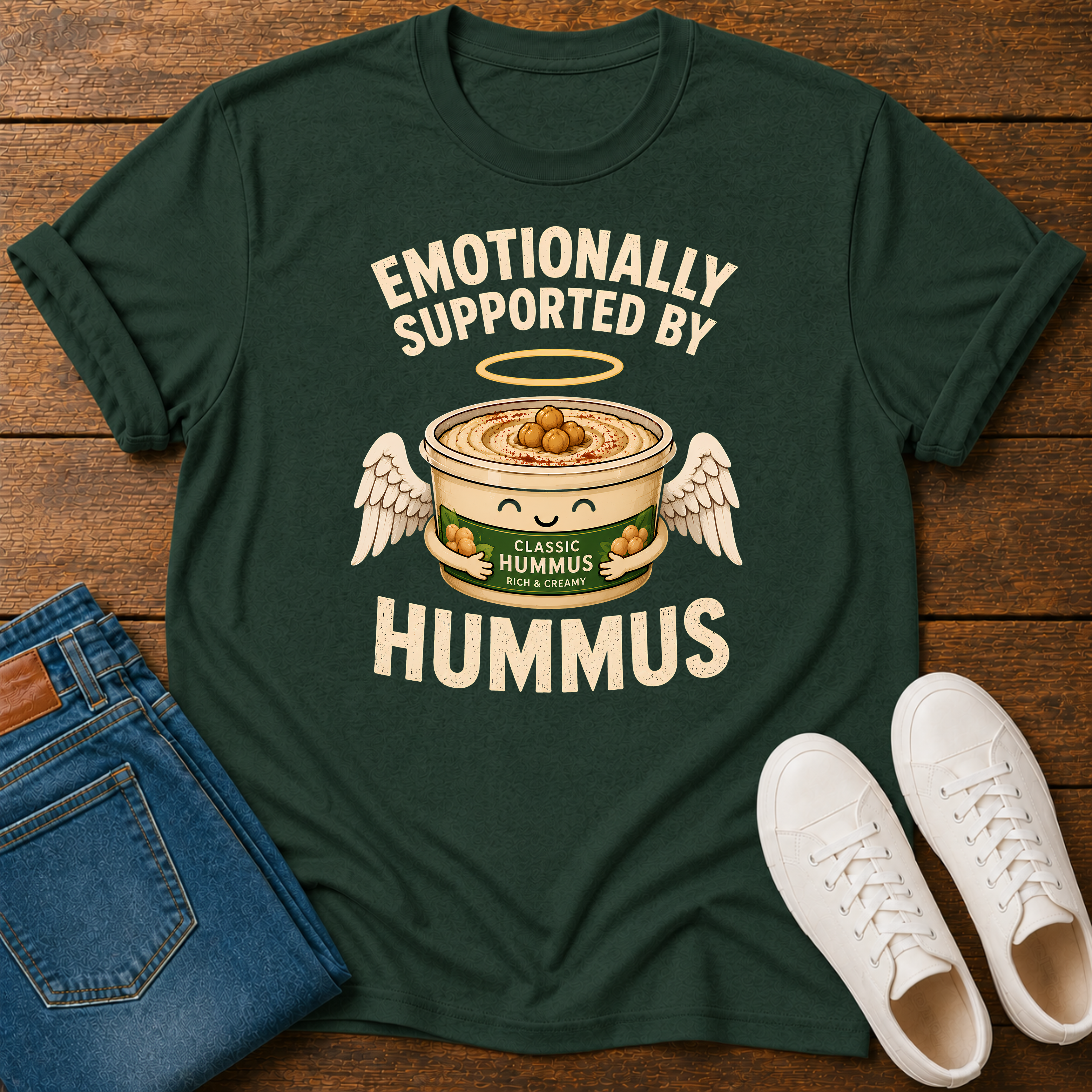 A dark green graphic t-shirt laid flat on a wooden surface, styled with blue jeans and white sneakers. The t-shirt features a humorous graphic of a cute, winged hummus bowl with the text 'EMOTIONALLY SUPPORTED BY HUMMUS'.