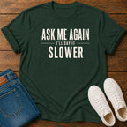 A dark green graphic t-shirt with the text 'ASK ME AGAIN I'LL SAY IT SLOWER' in white, styled in a flat lay on a wooden background with folded blue jeans and white sneakers.