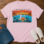 A light pink short-sleeve graphic T-shirt laid flat on a wooden surface, styled with folded blue jeans and white sneakers. The shirt has a large retro-style text graphic reading “I SURVIVED MY TRIP TO THE HYPOTHETICAL ISLAND” with palm trees and a cartoon face inside the lettering.