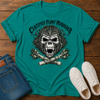 A teal short-sleeve graphic T-shirt laid flat on a wooden surface, styled with folded blue jeans and white sneakers. The shirt features a large black-and-white skull design with the text 'CERTIFIED PLANT MURDERER' above it.