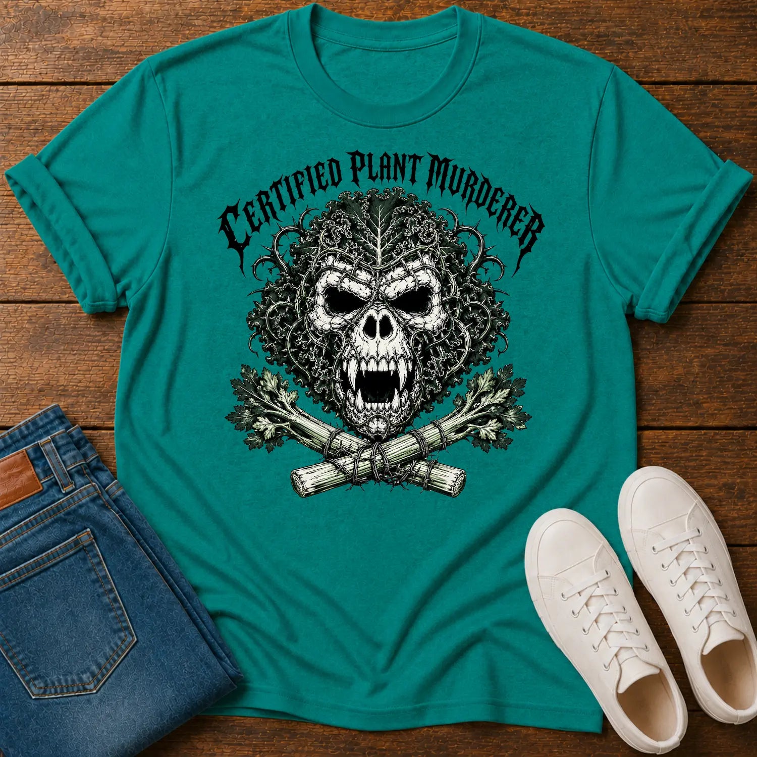 A teal short-sleeve graphic T-shirt laid flat on a wooden surface, styled with folded blue jeans and white sneakers. The shirt features a large black-and-white skull design with the text 'CERTIFIED PLANT MURDERER' above it.