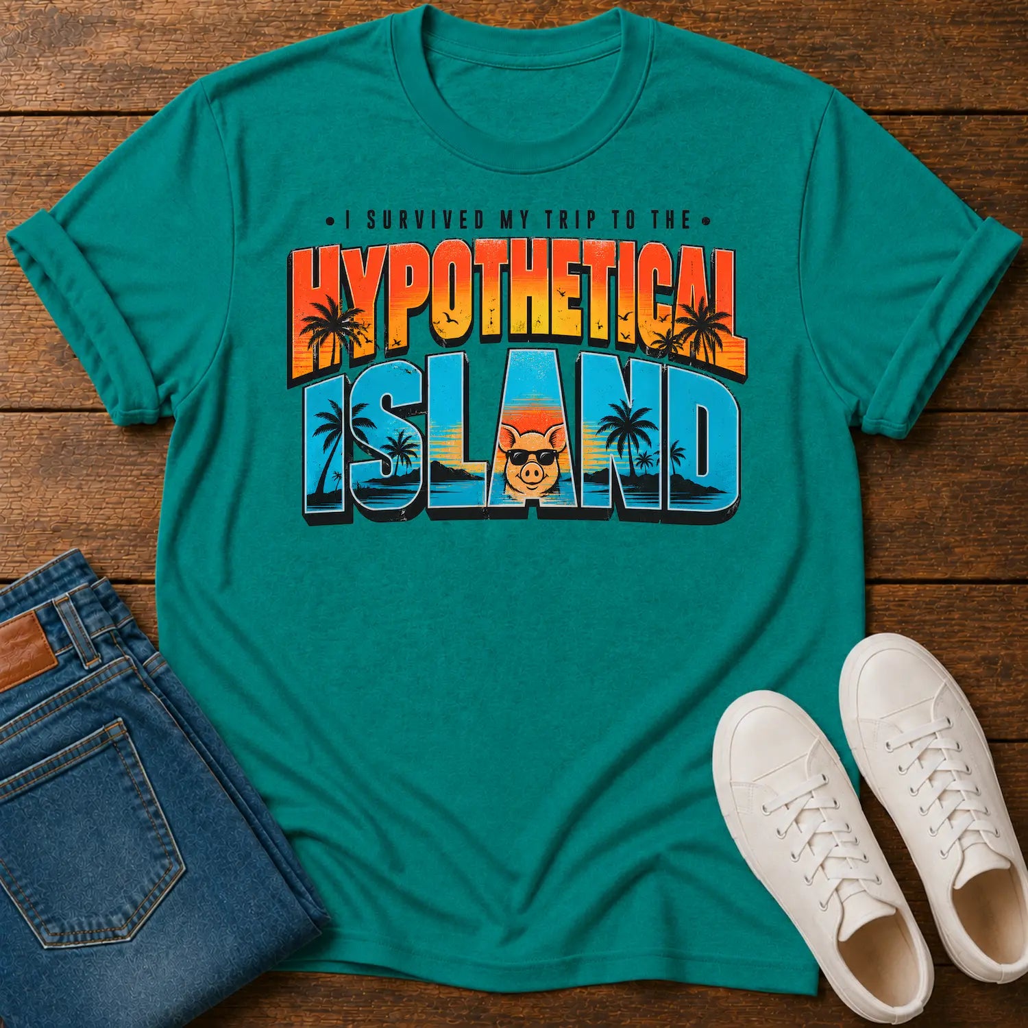 A jade short-sleeve graphic T-shirt laid flat on a wooden surface, styled with folded blue jeans and white sneakers. The shirt features a large retro-style text graphic reading “I SURVIVED MY TRIP TO THE HYPOTHETICAL ISLAND” with palm trees and a cartoon character wearing sunglasses.