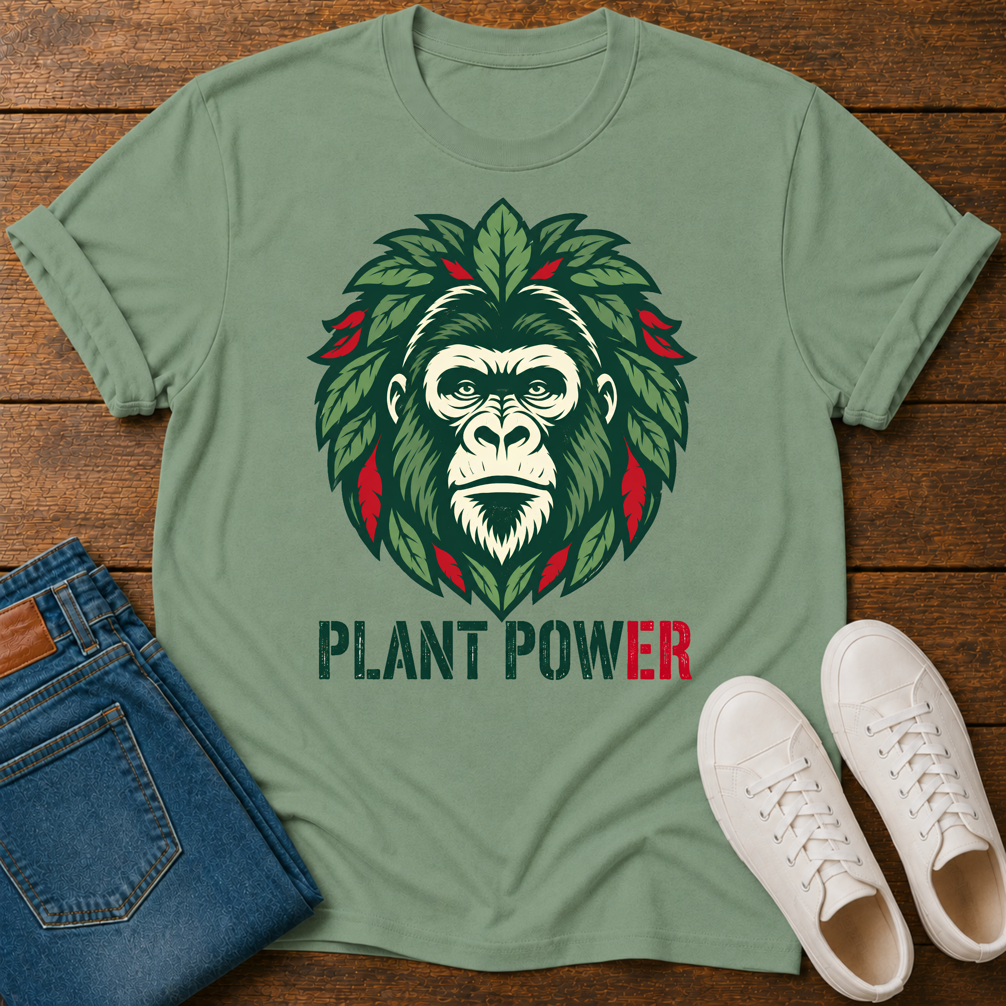 A sage green graphic t-shirt laid flat on a wooden surface, styled with folded blue jeans and white sneakers. The t-shirt features a graphic of a gorilla face surrounded by green leaves with red accents, and the text 'PLANT POWER' printed below.