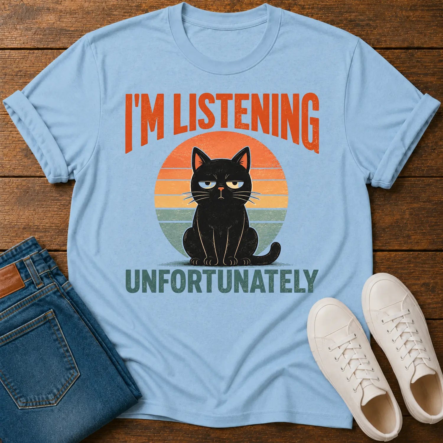A light blue graphic t-shirt laid flat on a dark wood background, styled with folded blue jeans and white sneakers. The t-shirt features a retro sunset graphic of a black cat with yellow eyes, with the text 'I'M LISTENING UNFORTUNATELY' in orange and dark green.