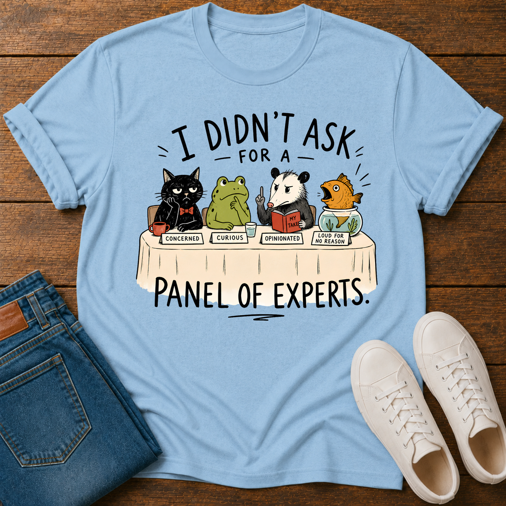 A light blue graphic t-shirt laid flat on a dark wood background, styled with folded blue jeans and a pair of white sneakers. The t-shirt features a cartoon panel of animals (a black cat, a green frog, a panda, and a fish) sitting at a table labeled 'I DIDN'T ASK FOR A PANEL OF EXPERTS.
