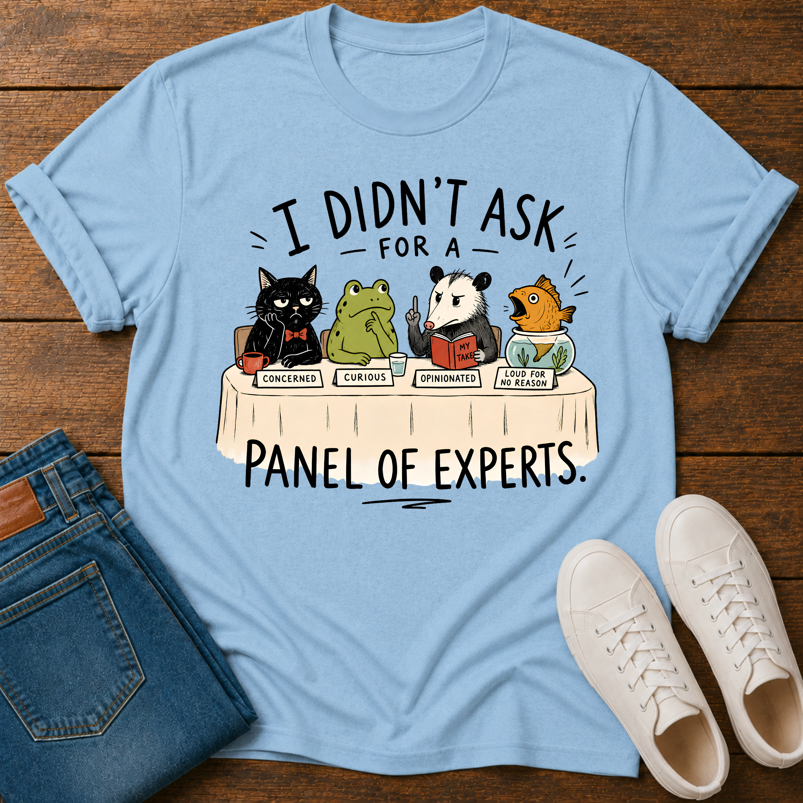 A light blue graphic t-shirt laid flat on a dark wood background, styled with folded blue jeans and a pair of white sneakers. The t-shirt features a cartoon panel of animals (a black cat, a green frog, a panda, and a fish) sitting at a table labeled 'I DIDN'T ASK FOR A PANEL OF EXPERTS.