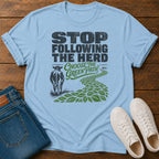 A light blue graphic t-shirt laid flat on a wooden surface, styled with folded blue jeans and white sneakers. The t-shirt features a graphic that reads 'STOP FOLLOWING THE HERD Choose the Green Path' with an illustration of a cow and a green leafy path.