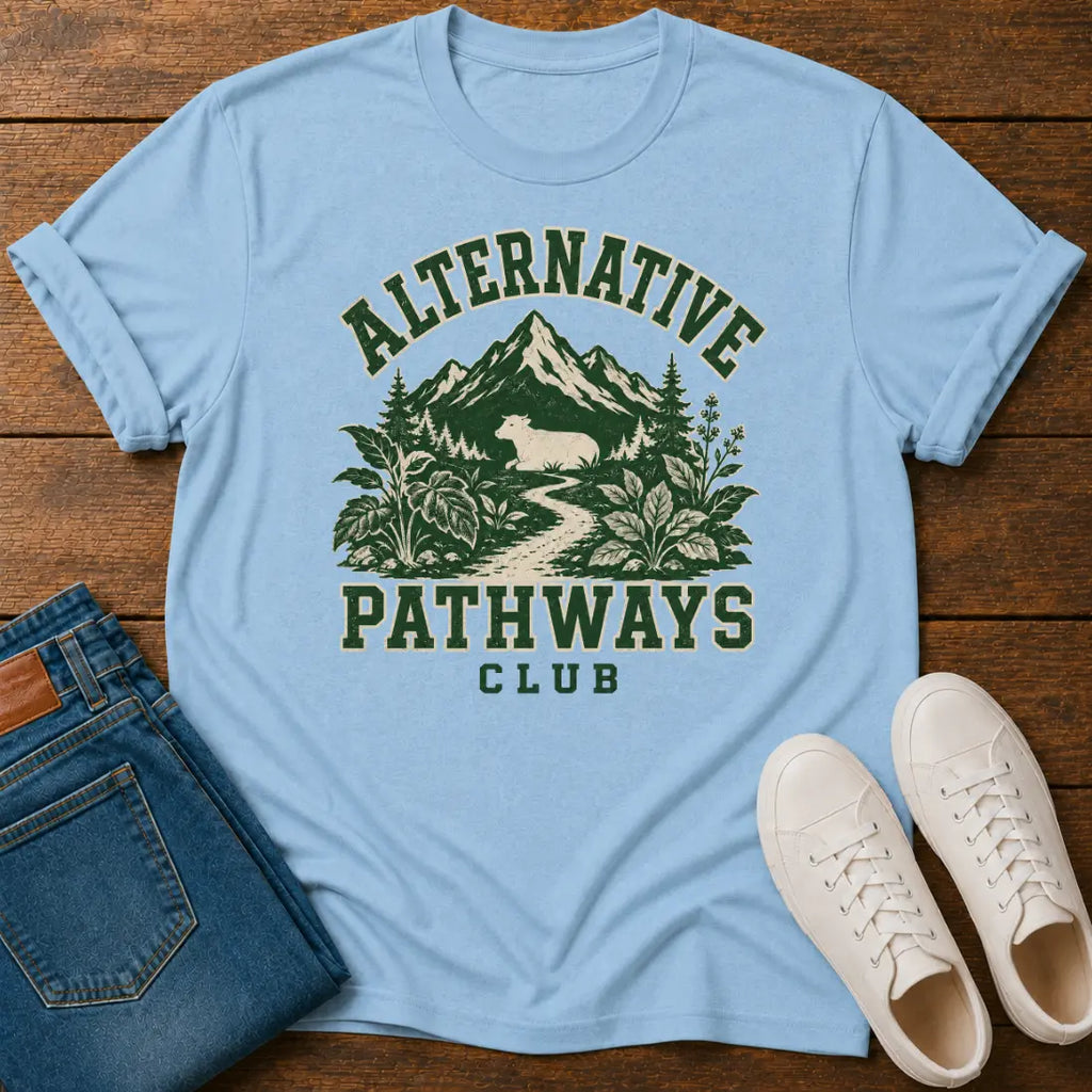 A light blue graphic t-shirt laid flat on a wooden surface, styled with folded blue jeans and white sneakers. The t-shirt features a dark green graphic of mountains, trees, and a deer with the text 'ALTERNATIVE PATHWAYS CLUB'.