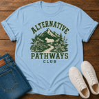 A light blue graphic t-shirt laid flat on a wooden surface, styled with folded blue jeans and white sneakers. The t-shirt features a dark green graphic of mountains, trees, and a deer with the text 'ALTERNATIVE PATHWAYS CLUB'.