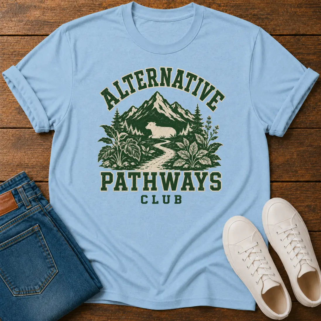 A light blue graphic t-shirt laid flat on a wooden surface, styled with folded blue jeans and white sneakers. The t-shirt features a dark green graphic of mountains, trees, and a deer with the text 'ALTERNATIVE PATHWAYS CLUB'.