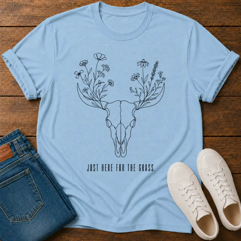 A light blue graphic t-shirt laid flat on a wooden surface, styled with folded blue jeans and white sneakers. The t-shirt features a black line art design of a deer skull with wildflowers for antlers and the text 'JUST HERE FOR THE GRASS.