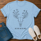 A light blue graphic t-shirt laid flat on a wooden surface, styled with folded blue jeans and white sneakers. The t-shirt features a black line art design of a deer skull with wildflowers for antlers and the text 'JUST HERE FOR THE GRASS.