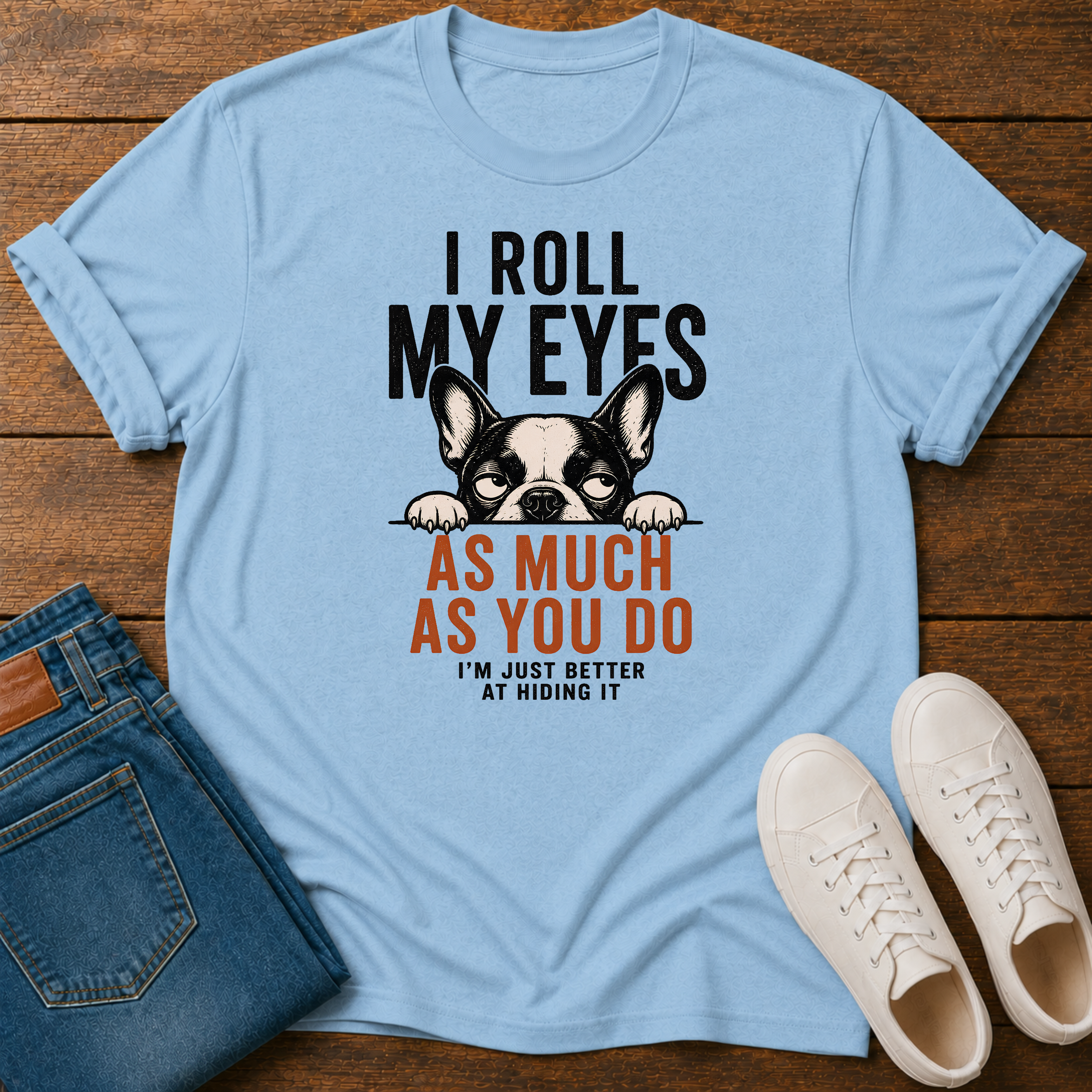 A light blue graphic t-shirt laid flat on a wooden surface, styled with folded blue jeans and white sneakers. The t-shirt features a black and white French Bulldog graphic with the text 'I ROLL MY EYES AS MUCH AS YOU DO I'M JUST BETTER AT HIDING IT'.
