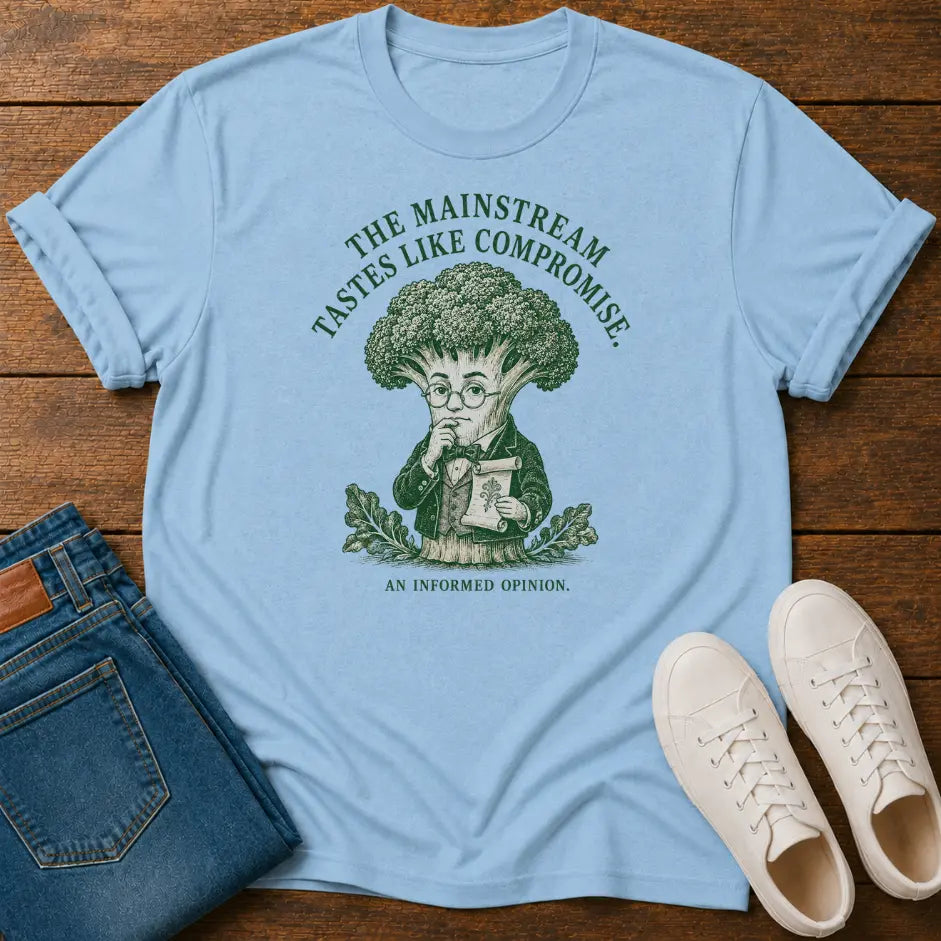 A light blue graphic t-shirt laid flat on a wooden surface, styled with folded blue jeans and white sneakers. The t-shirt features a green graphic of a broccoli-headed character reading a book, with the text 'THE MAINSTREAM TASTES LIKE COMPROMISE.