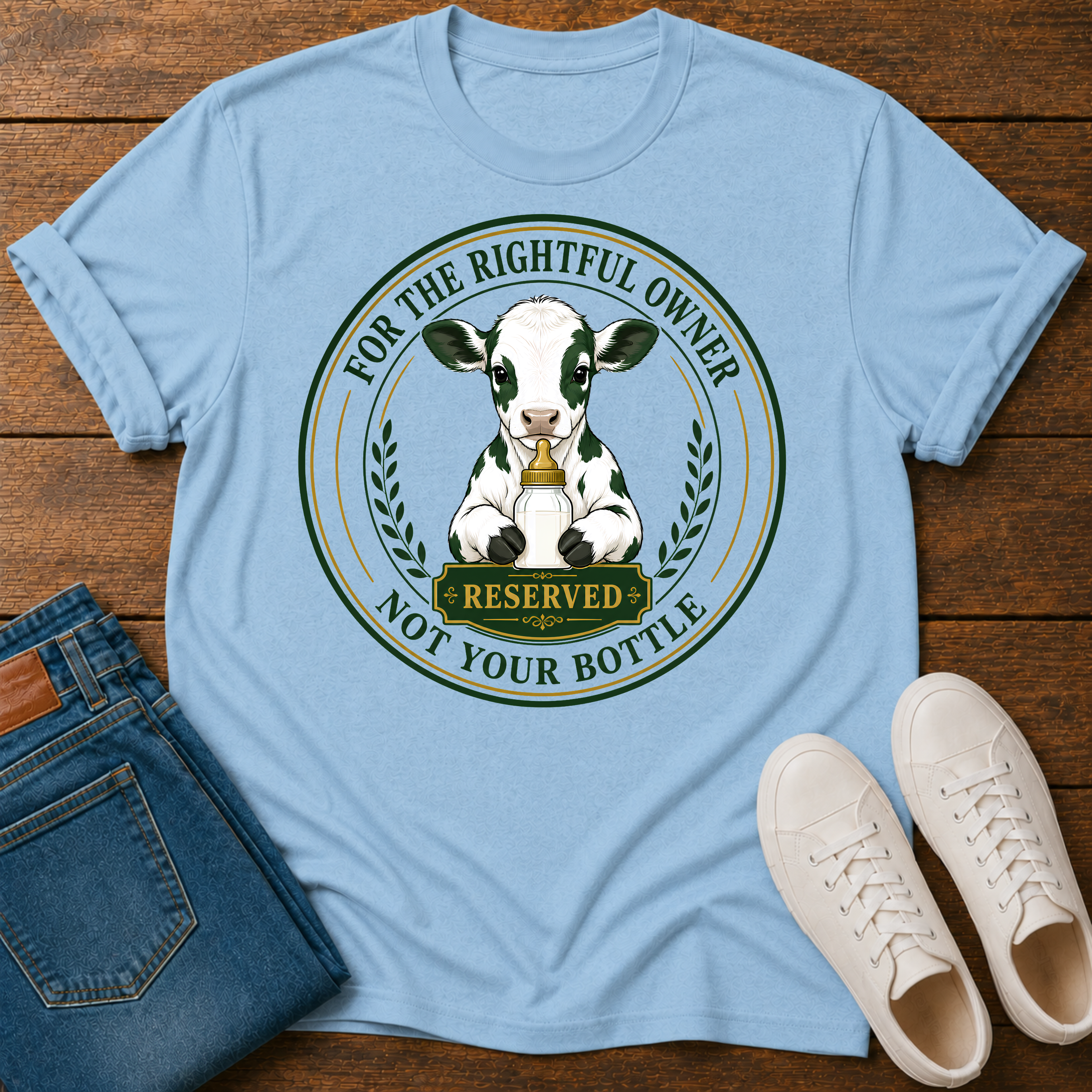 A light blue graphic t-shirt laid flat on a wooden surface, styled with folded blue jeans and white sneakers. The t-shirt features a circular graphic of a black-and-white cow with a gold bell and the text 'FOR THE RIGHTFUL OWNER NOT YOUR BOTTLE' and 'RESERVED'.