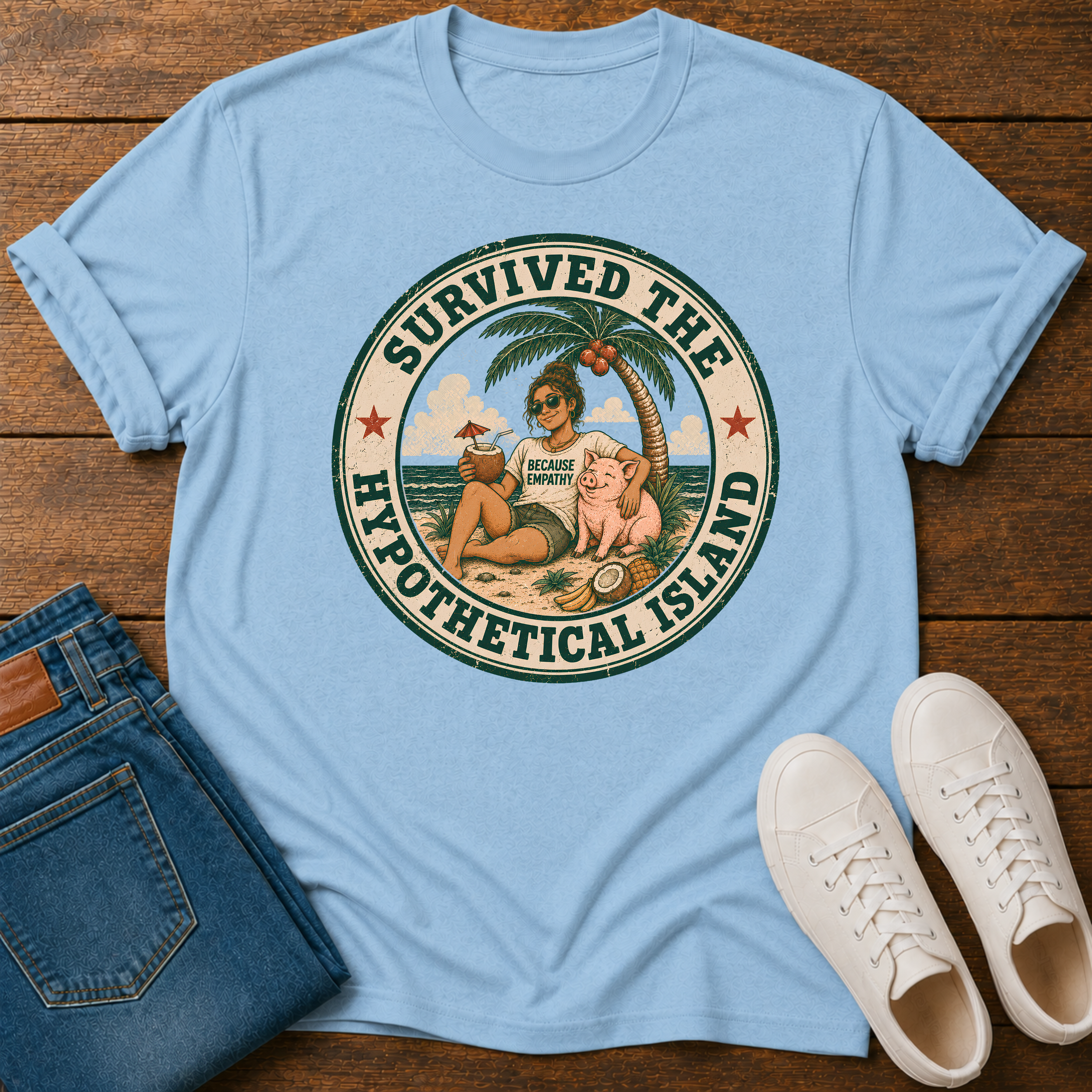 A light blue crewneck t-shirt laid flat on a dark wood background, styled with folded blue jeans and white sneakers. The t-shirt features a circular graphic print with the text 'SURVIVED THE HYPOTHETICAL ISLAND' and an illustration of a man wearing a 'HYPOTHETICAL MAN CLOTHING' shirt with a coconut drink and a pig.