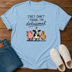 A light blue graphic t-shirt laid flat on a dark wood background, styled with folded blue jeans and a pair of white sneakers. The t-shirt features a humorous graphic that reads 'THEY DON'T THINK I'M delusional' above an illustration of a pink pig, a black-and-white cow, and a yellow chicken.