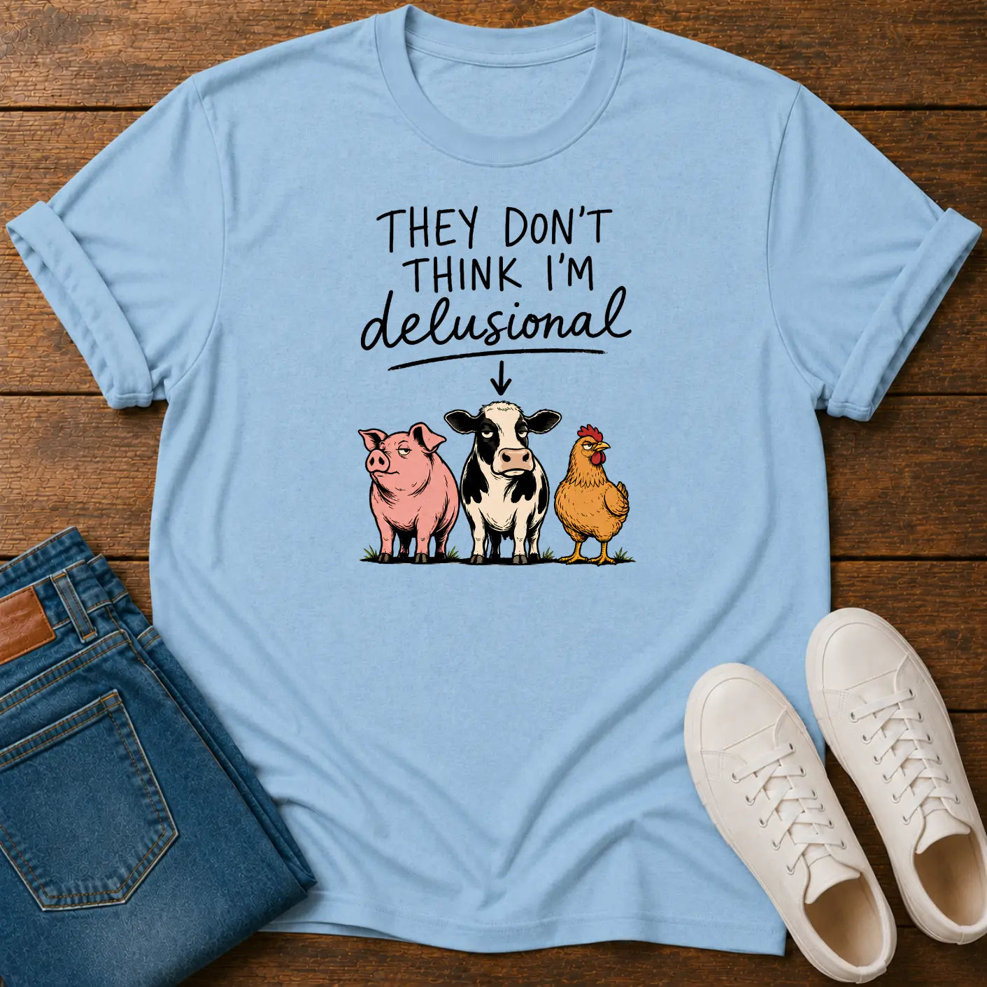 A light blue graphic t-shirt laid flat on a dark wood background, styled with folded blue jeans and a pair of white sneakers. The t-shirt features a humorous graphic that reads 'THEY DON'T THINK I'M delusional' above an illustration of a pink pig, a black-and-white cow, and a yellow chicken.
