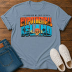 A light blue graphic t-shirt laid flat on a wooden surface, styled with folded blue jeans and white sneakers. The t-shirt features a colorful retro-style graphic that reads 'I SURVIVED MY TRIP TO THE HYPOTHETICAL ISLAND' with palm trees and a cartoon character.