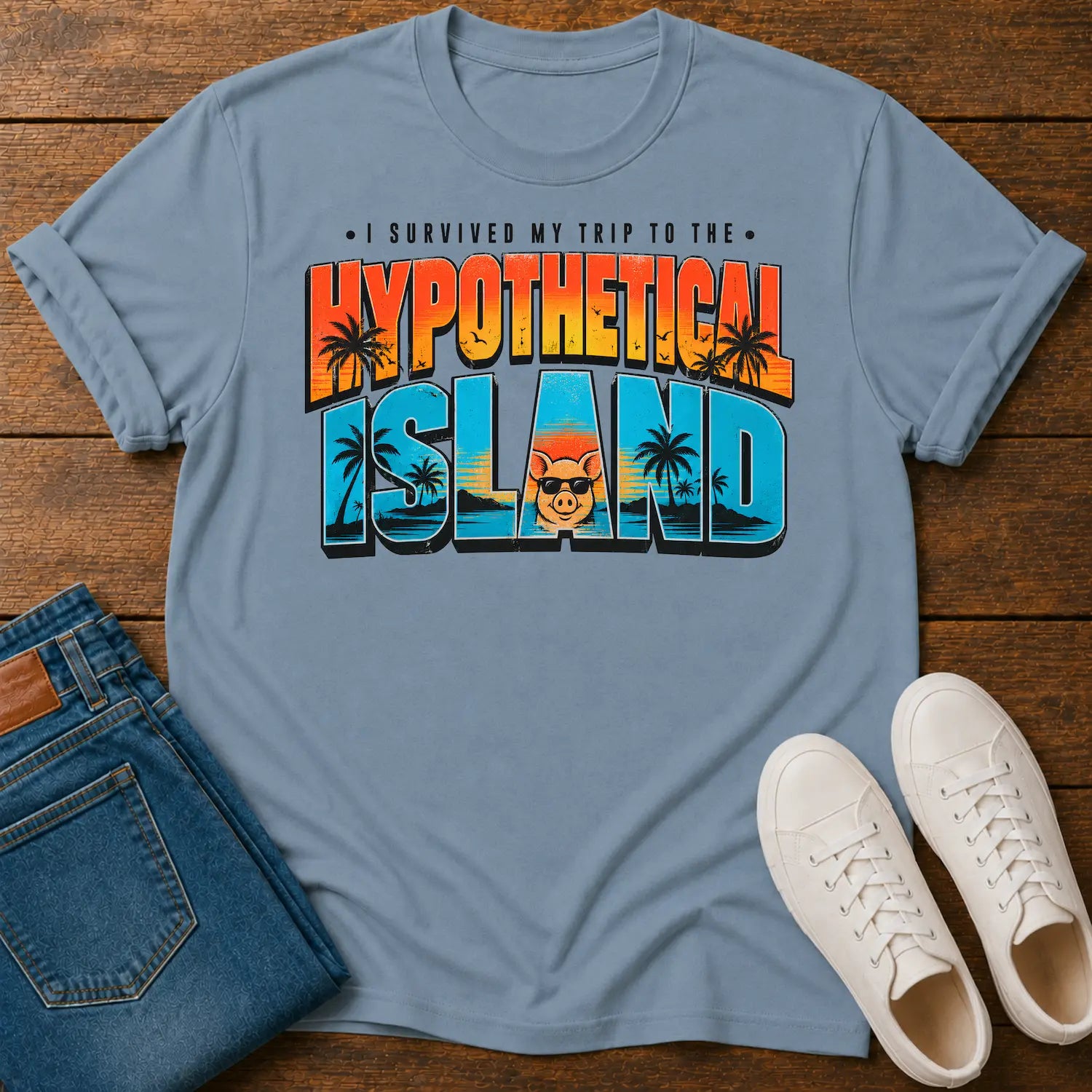 A light blue graphic t-shirt laid flat on a wooden surface, styled with folded blue jeans and white sneakers. The t-shirt features a colorful retro-style graphic that reads 'I SURVIVED MY TRIP TO THE HYPOTHETICAL ISLAND' with palm trees and a cartoon character.