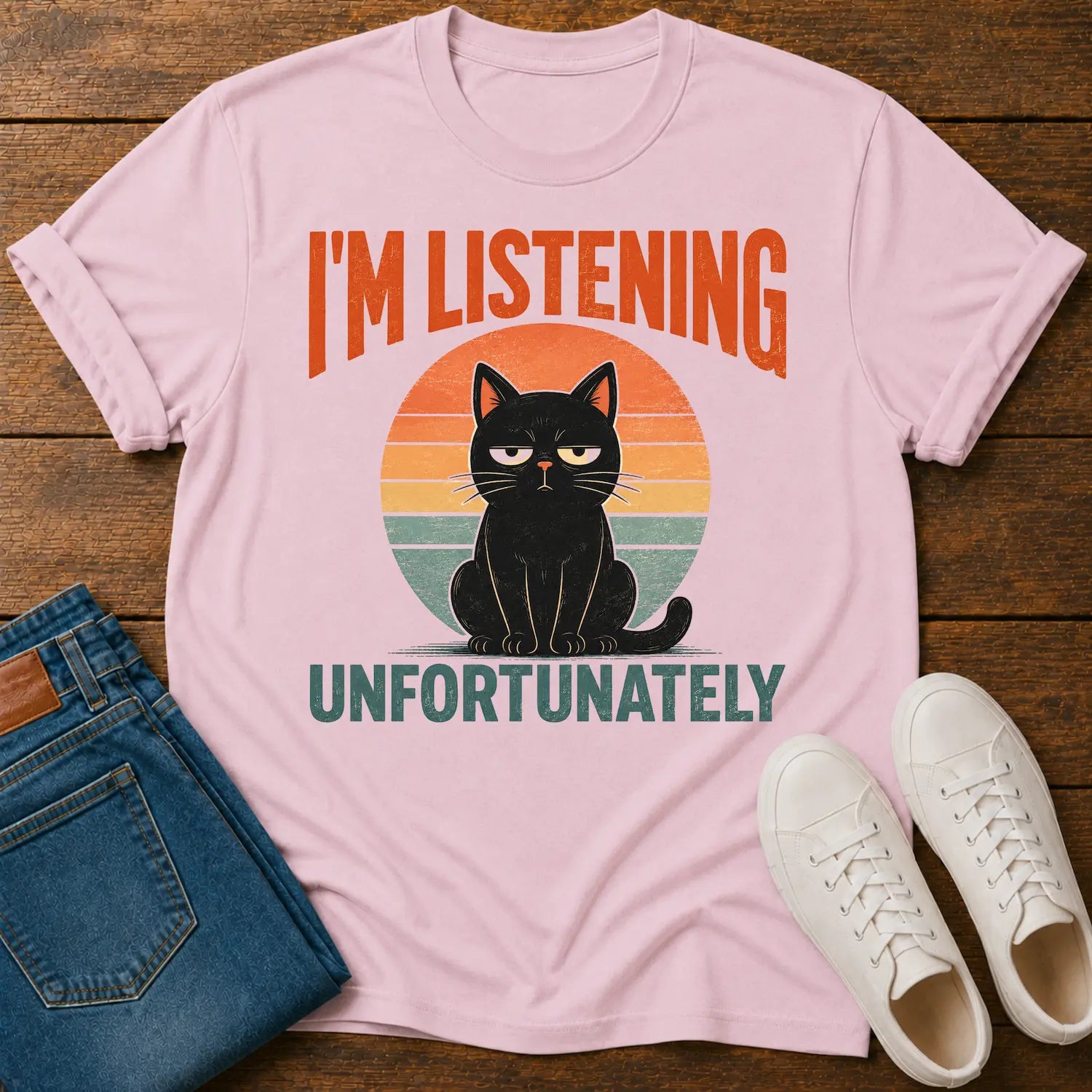 A light pink graphic t-shirt laid flat on a rustic wooden background, styled with folded blue jeans and white sneakers. The t-shirt features a retro design with a black cat sitting in front of a sunset circle and the text 'I'M LISTENING UNFORTUNATELY'.