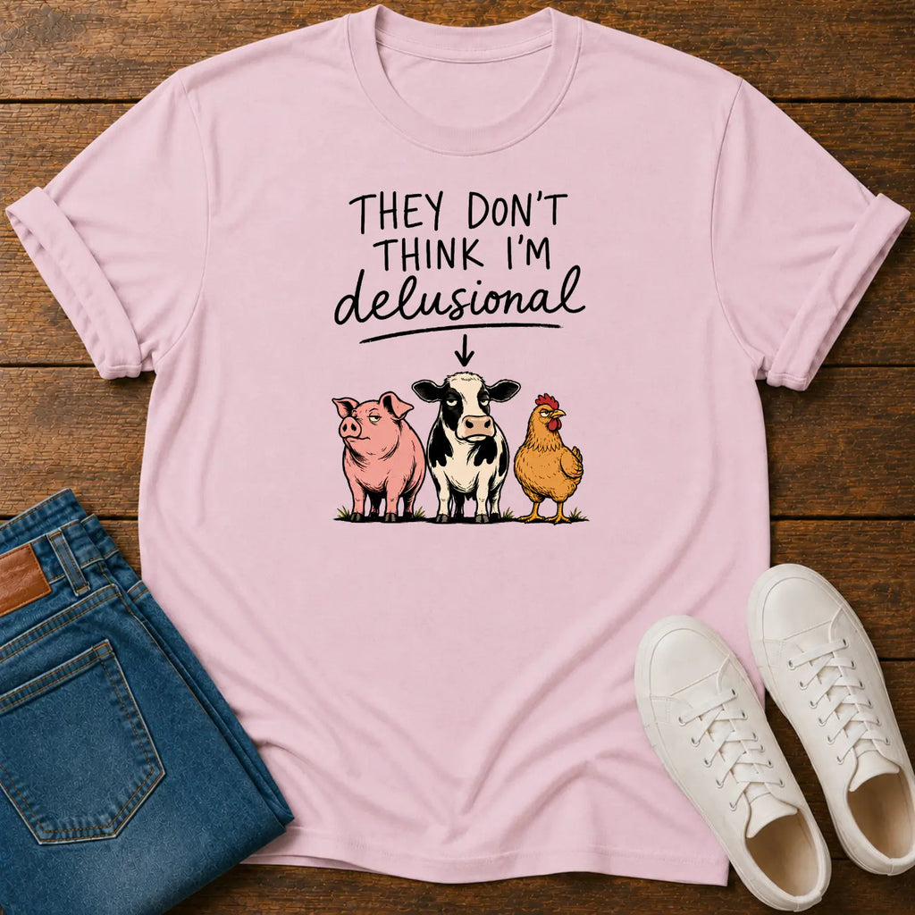A light pink graphic t-shirt laid flat on a dark wood background, styled with folded blue jeans and white sneakers. The t-shirt features the text 'THEY DON'T THINK I'M delusional' above an illustration of a pink pig, a black-and-white cow, and a yellow chicken.